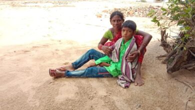 Mother sits by the riverbank holding her sick son; questions raised about the healthcare system.