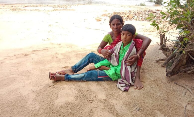 Mother sits by the riverbank holding her sick son; questions raised about the healthcare system.